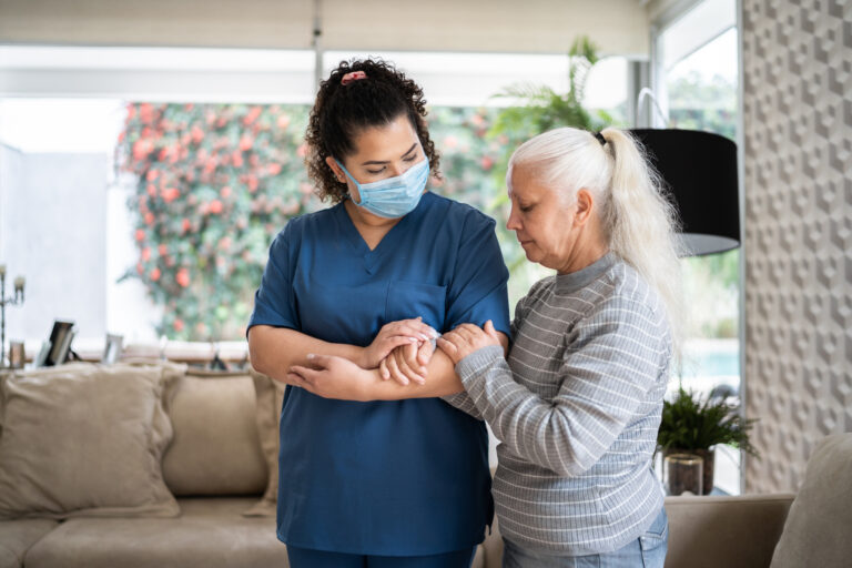 Home Caregiver Helping Senior Woman Walking At Home Wearing Protecive Face Mask